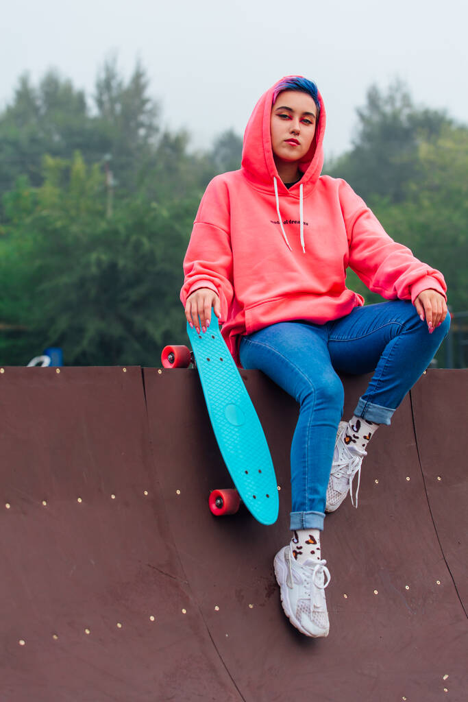 Estilo de vida de verano imagen de moda chica bonita vestida con sudadera con capucha rosa y vaqueros sentados junto a la pista de skate con su patín de plástico azul. - Foto, imagen