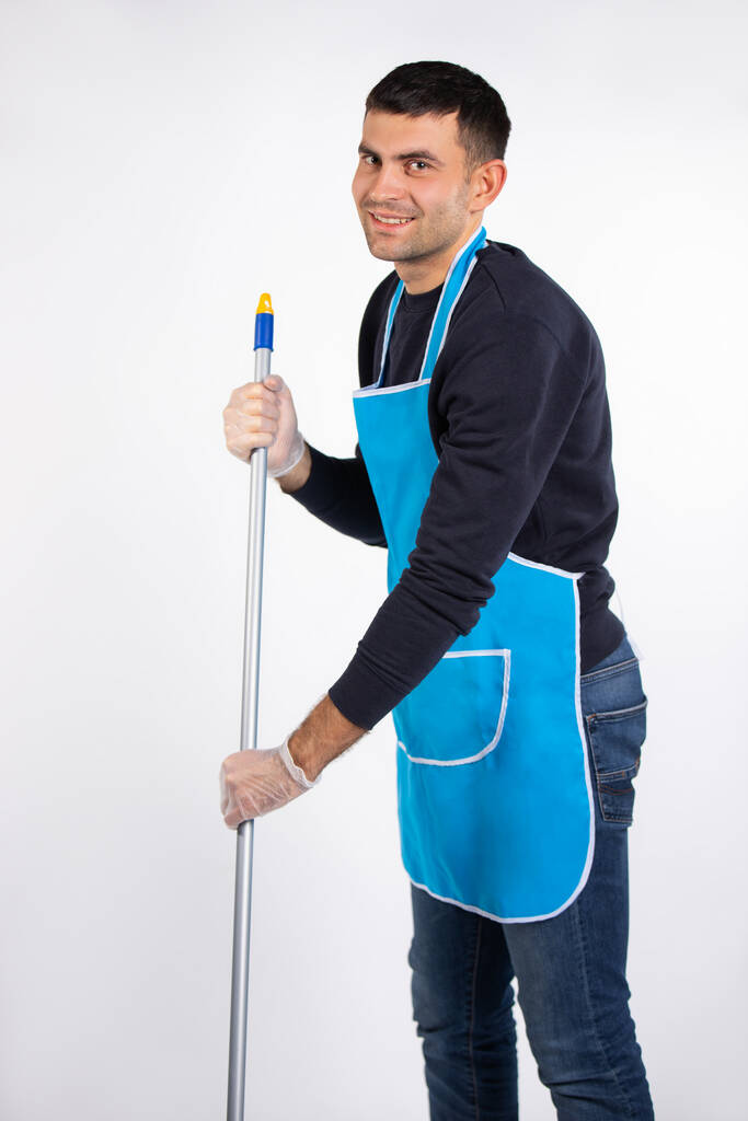 Ένας άντρας με ποδιά και γάντια σκουπίζει το πάτωμα με μια σφουγγαρίστρα με χαμόγελο. Καταπολέμηση στερεοτύπων φύλου. Φωτογραφία σε λευκό φόντο. Υψηλής ποιότητας φωτογραφία - Φωτογραφία, εικόνα