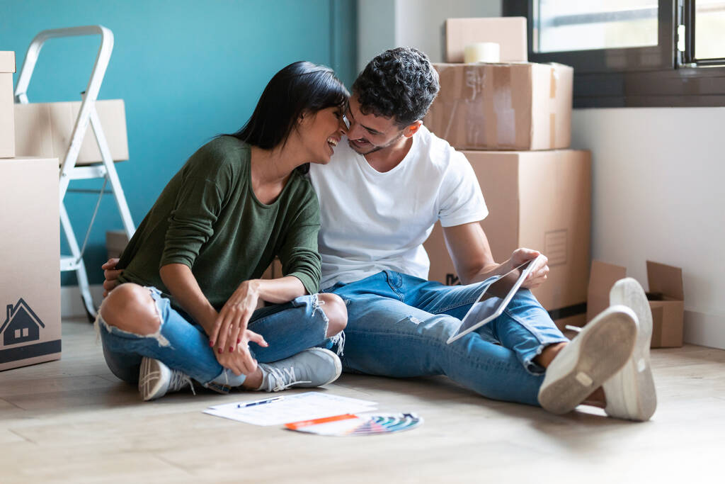 Shot χαριτωμένο ζευγάρι φιλιούνται, ενώ ψάχνει ιδέες για να διακοσμήσετε το νέο διαμέρισμα στο ψηφιακό tablet κάθεται στο πάτωμα. - Φωτογραφία, εικόνα