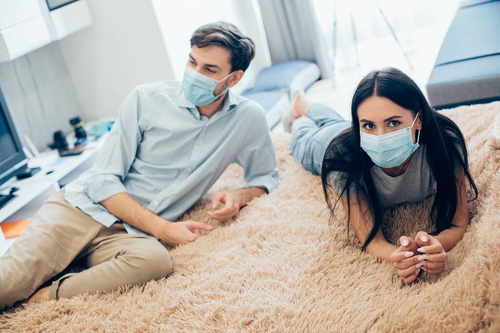 Joven hombre y mujer en máscaras médicas tumbados en la cama en su piso mientras se relaja en el auto-aislamiento - Foto, Imagen