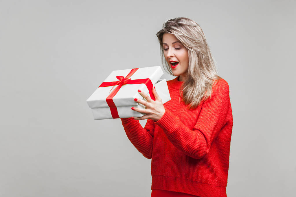 Portrait of surprised beautiful blonde woman with red lipstick in bright casual sweater, looking inside gift box with amazement, unboxing present. indoor studio shot isolated on gray background - Photo, Image