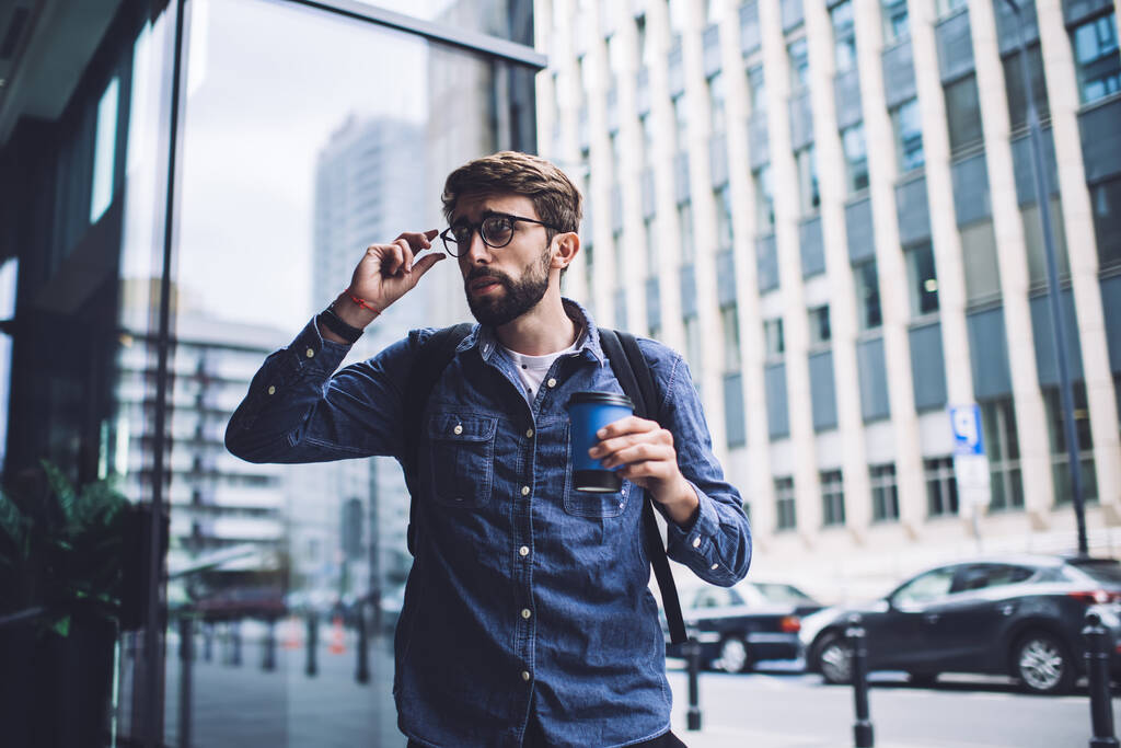 Χαρούμενος γενειοφόρος άνδρας φορώντας casual ρούχα και γυαλιά που στέκεται με ζεστό καφέ και κοιτάζοντας μακριά προσεκτικά κοντά στο σύγχρονο γυάλινο κτίριο - Φωτογραφία, εικόνα