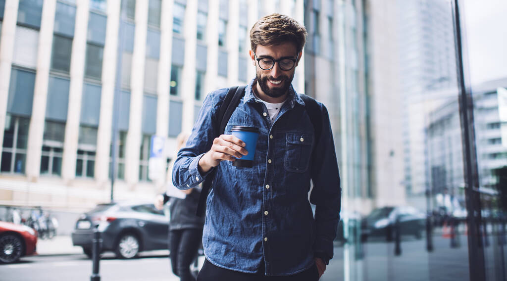 Pozitív szakállas férfi hipster visel alkalmi ruhát áll az utcán, és néz le, miközben iszik kávét közelében kortárs üveg épületek - Fotó, kép