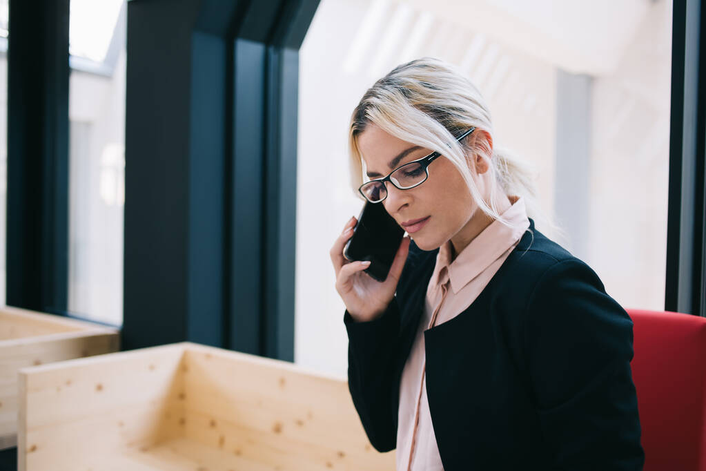 Ciddi stil sahibi, siyah takım elbiseli ve gözlüklü kadın girişimci modern çalışma alanının penceresinin yanında otururken telefonla konuşuyor. - Fotoğraf, Görsel