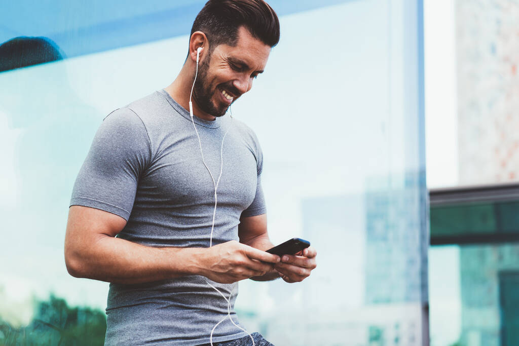 Cheerful Caucasian bodybuilder in electronic earphones listening received audio message during positive friendly chatting via smartphone application, happy male athlete 30s sending mobile sms - Photo, Image