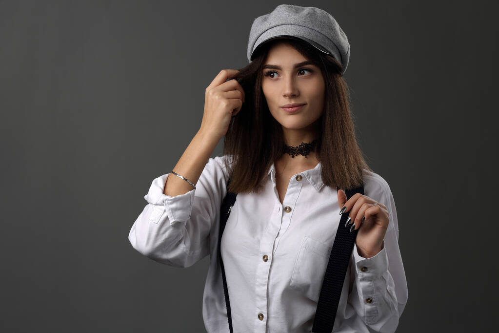 Gri arka planda siyah pantolon askısı olan beyaz ve şapkalı güzel bir kızın portresi. - Fotoğraf, Görsel