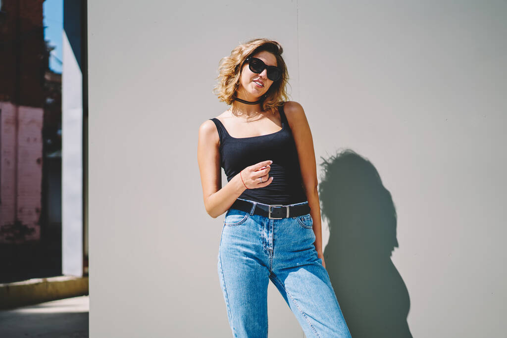 Молодая улыбающаяся женщина с каштановыми волосами в солнцезащитных очках в джинсах и удушающих позах стоя на сером фоне на солнечной улице - Фото, изображение