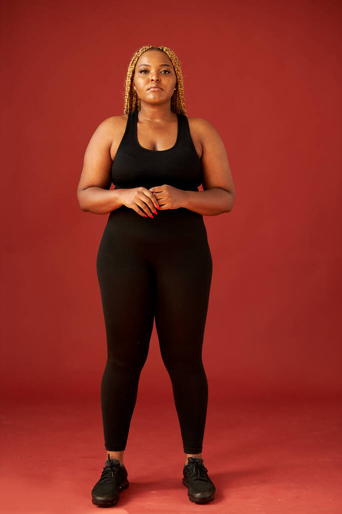 zelfverzekerde vet Afrikaanse vrouw in zwart sportkleding geïsoleerd op rode achtergrond - Foto, afbeelding