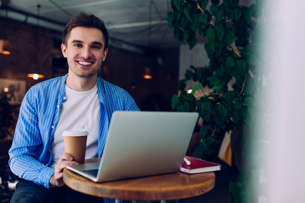 Contenu pigiste masculin en tenue décontractée assis à une petite table ronde avec ordinateur portable ouvert tout en prenant le café au café - Photo, image