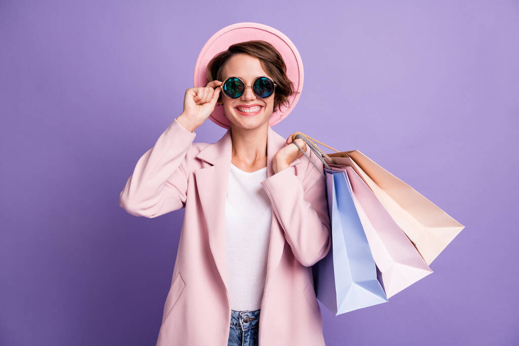 Photo of sweet pretty optimistic bob hair young woman carry shop bags wear glasses pink cap coat isolated on purple color background - Photo, Image