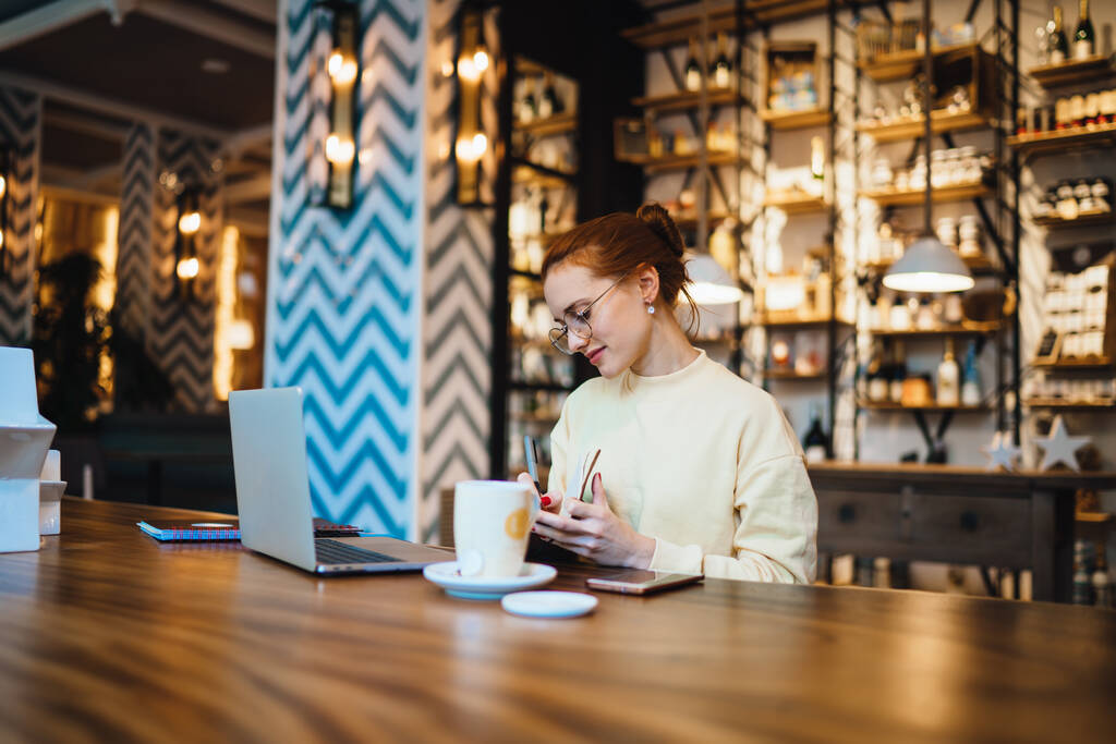 Свободная красивая девушка в повседневной одежде и очках сидит за столом с кружкой чая и мобильной записью в блокноте, просматривая ноутбук в кафе в Барселоне - Фото, изображение