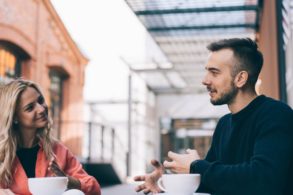 Счастливая прекрасная леди и бородатый мужчина в повседневной одежде улыбаются и смотрят друг на друга, наслаждаясь кофе на террасе кафе в солнечный день - Фото, изображение