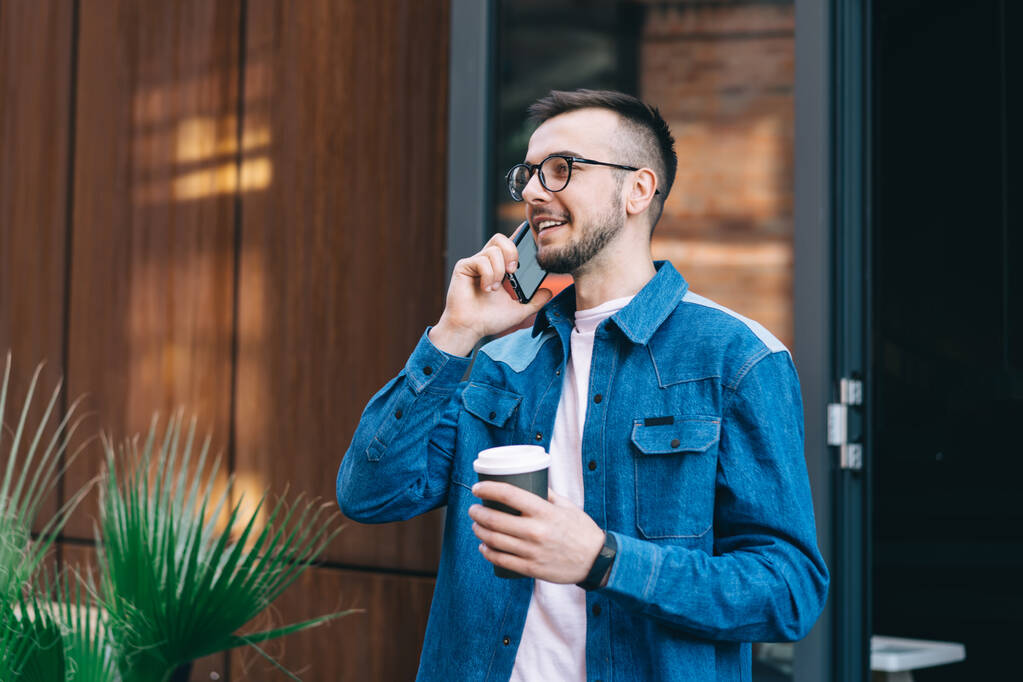 Sorrindo hipster masculino em roupas casuais e óculos de pé com xícara de café e se comunicando no telefone celular na rua - Foto, Imagem