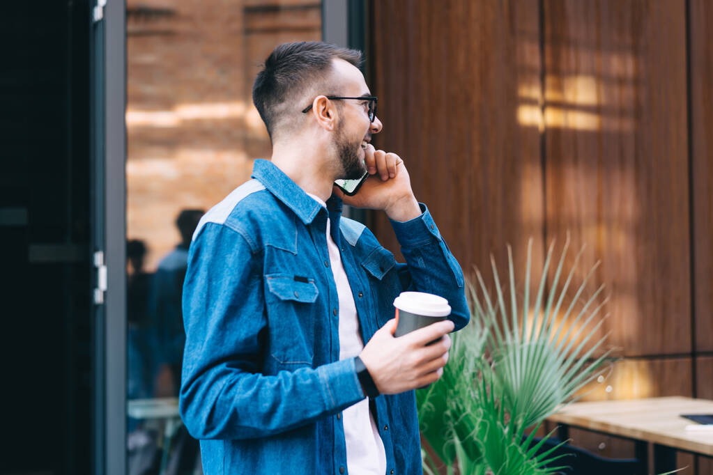 Side view of bearded student in casual clothing holding tea in paper cup and laughing while talking on smartphone near urban cafe - Photo, Image