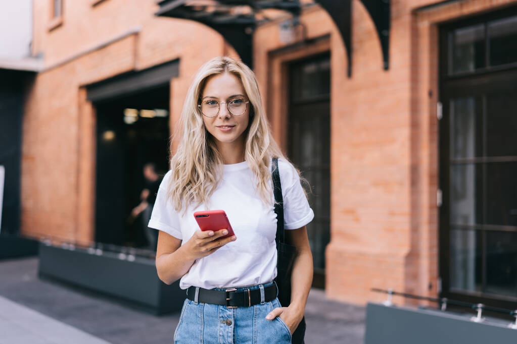 Glückliche junge blonde Frau in lässiger Kleidung und Brille surft auf dem Smartphone im Internet, während sie mit der Hand in der Tasche auf der Straße steht und in die Kamera lächelt - Foto, Bild