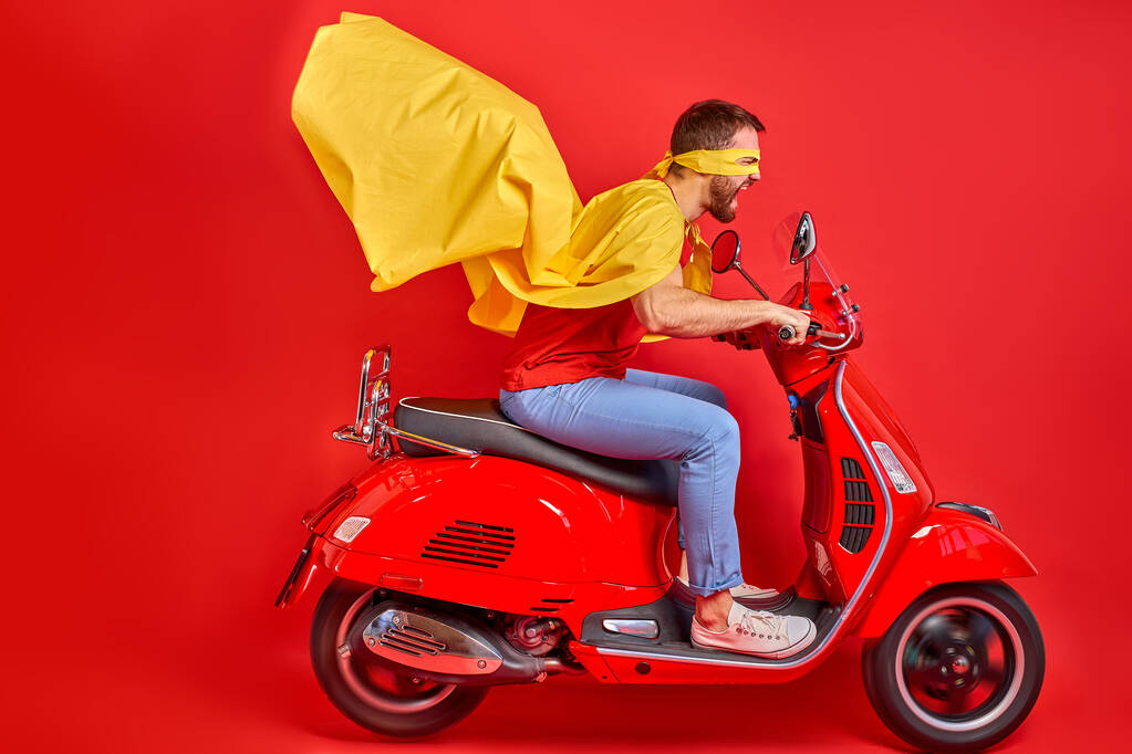 Сумасшедший парень водит мопед в плаще и маске, спеша на дорожную вечеринку в роли супергероев - Фото, изображение