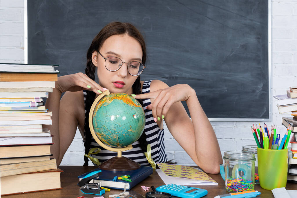 Durante una lección de geografía, una adolescente observa el mundo y planea tranquilamente sus viajes de verano. Una joven con gafas se sienta en un escritorio de la escuela. - Foto, Imagen