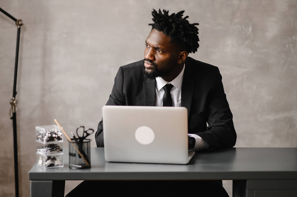  Όμορφος Αφροαμερικάνος επιχειρηματίας με κλασική στολή χρησιμοποιεί φορητό υπολογιστή και χαμόγελα ενώ εργάζεται στο γραφείο - Φωτογραφία, εικόνα