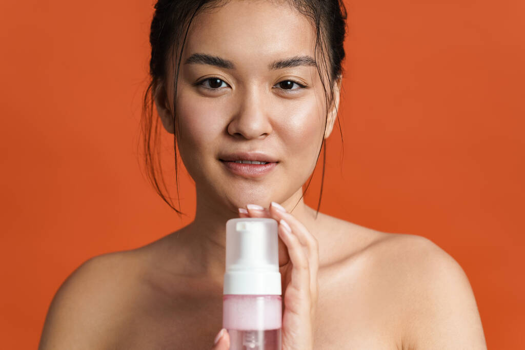 Imagen de la chica asiática sin camisa complacida posando con espuma limpiadora aislada sobre fondo naranja - Foto, Imagen