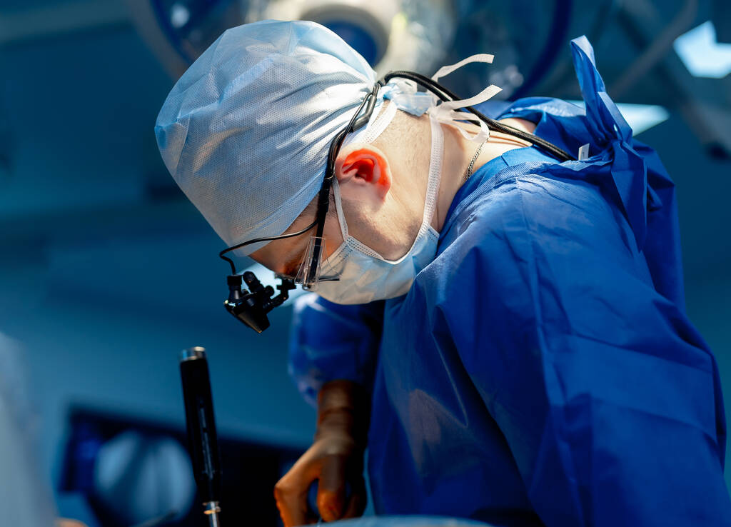 Neurochirurg in blauwe scrubs en chirurgische bril biedt operatie in up-to-date operatiekamer met moderne apparatuur. - Foto, afbeelding