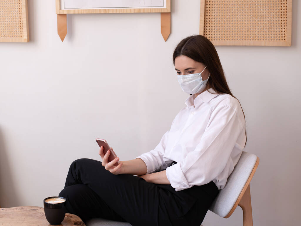 Egy fiatal nő egy kávézóban orvosi maszkot visel. Nézd a telefont.. - Fotó, kép