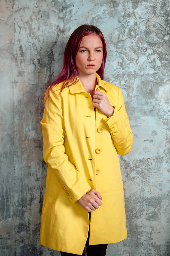 Hermosa mujer con cabello rosa vestida con impermeable amarillo tocando su cara y posando en el interior cerca de la pared de hormigón. - Foto, imagen