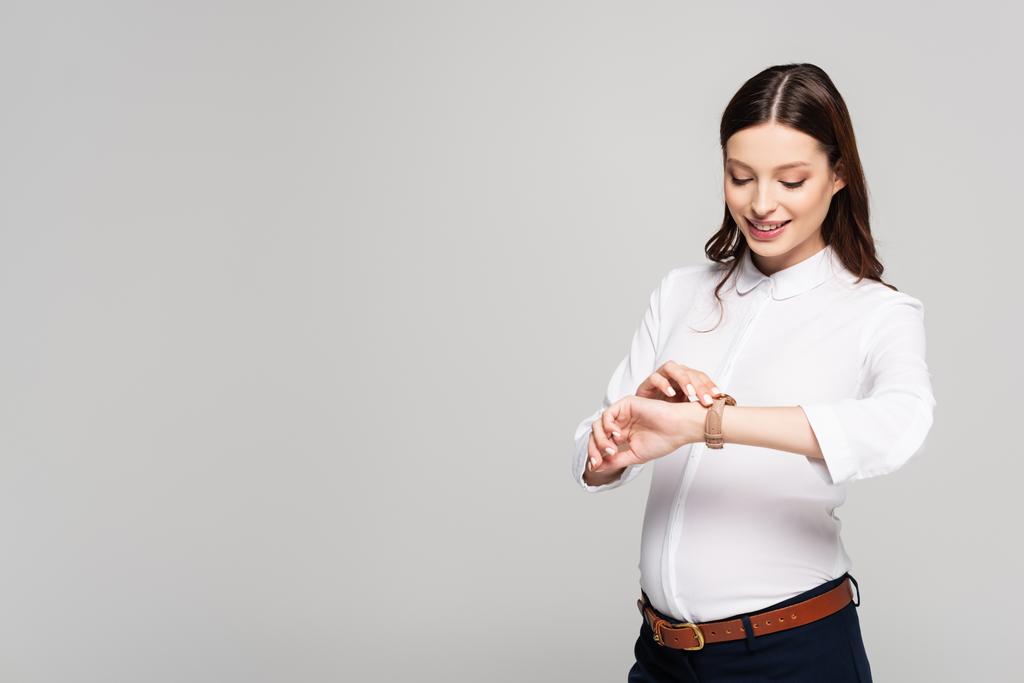 sorridente giovane donna d'affari incinta guardando orologio da polso isolato su grigio - Foto, immagini