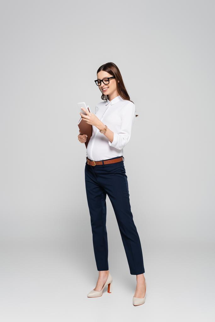 улыбающаяся молодая беременная деловая женщина в очках с папкой и смартфоном, изолированными на сером - Фото, изображение