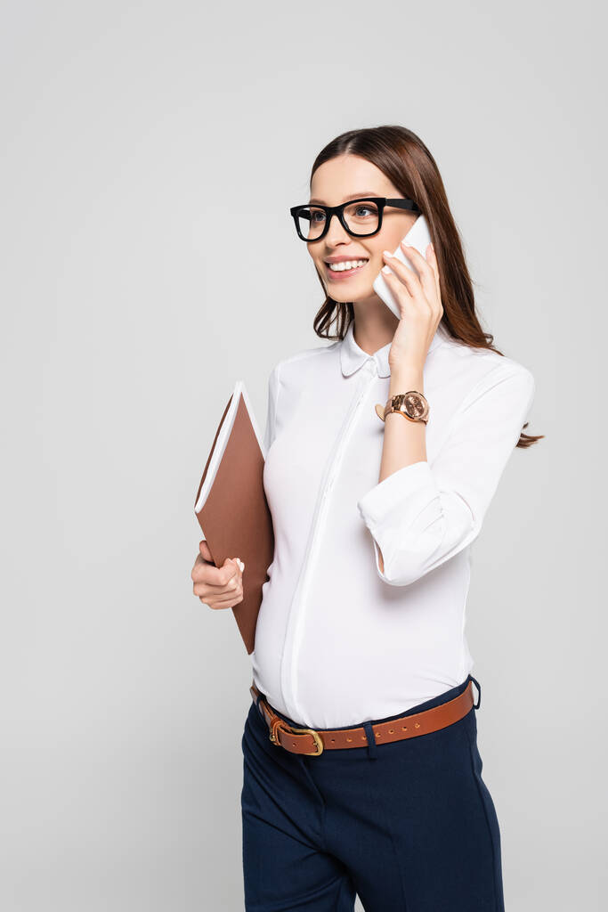 sonriente joven embarazada mujer de negocios en gafas con carpeta hablando en smartphone aislado en gris - Foto, Imagen