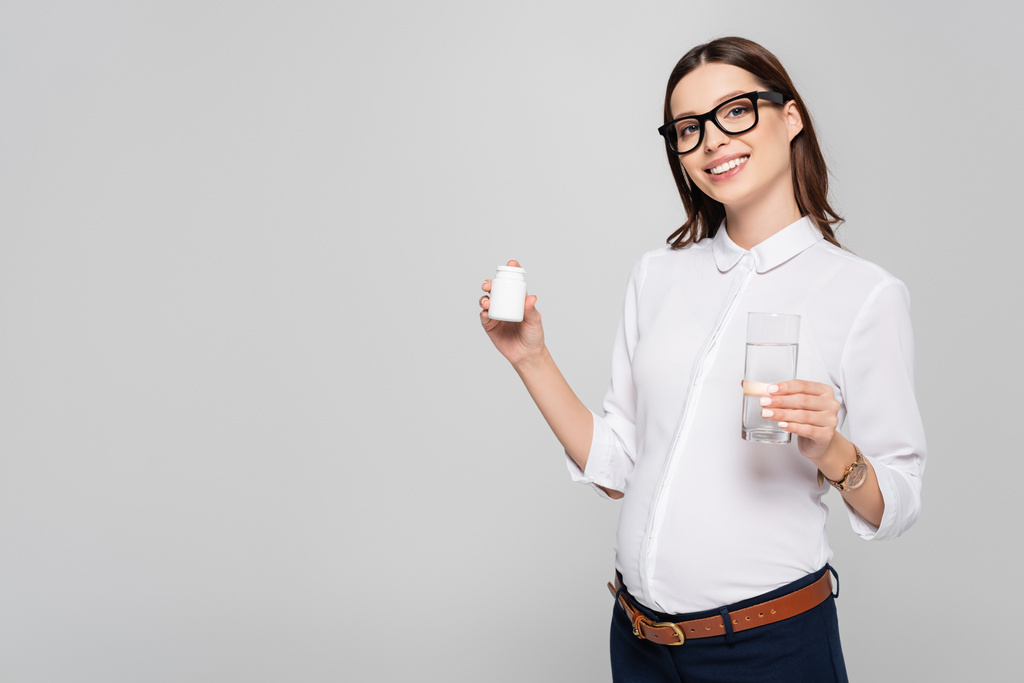 улыбающаяся молодая беременная деловая женщина в очках со стаканом воды и витаминами для беременных, изолированных на сером - Фото, изображение