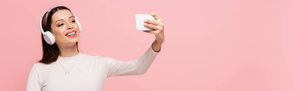 szczęśliwy młody ładna kobieta w słuchawki biorąc selfie na smartfon odizolowany na różowy, baner - Zdjęcie, obraz