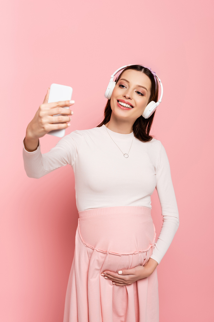 щаслива молода красива вагітна жінка в навушниках за допомогою смартфона ізольована на рожевому
 - Фото, зображення