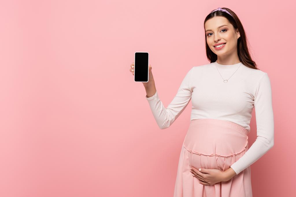 Ευτυχισμένη νεαρή όμορφη έγκυος γυναίκα δείχνει smartphone απομονώνονται σε ροζ - Φωτογραφία, εικόνα