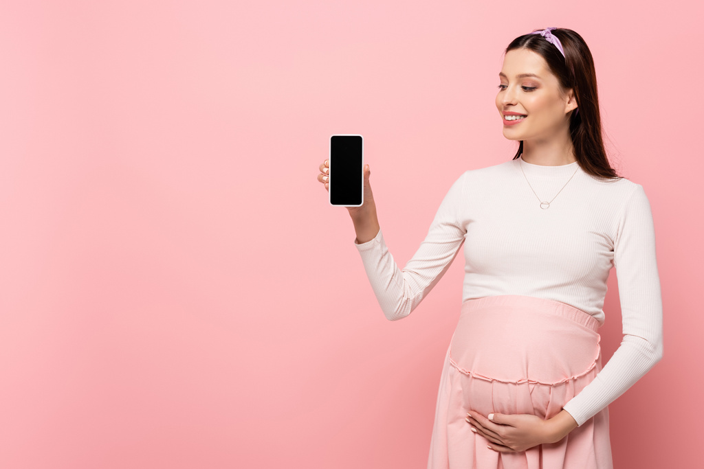Mutlu, genç, güzel, hamile bir kadın pembe üzerine izole akıllı telefon gösteriyor. - Fotoğraf, Görsel