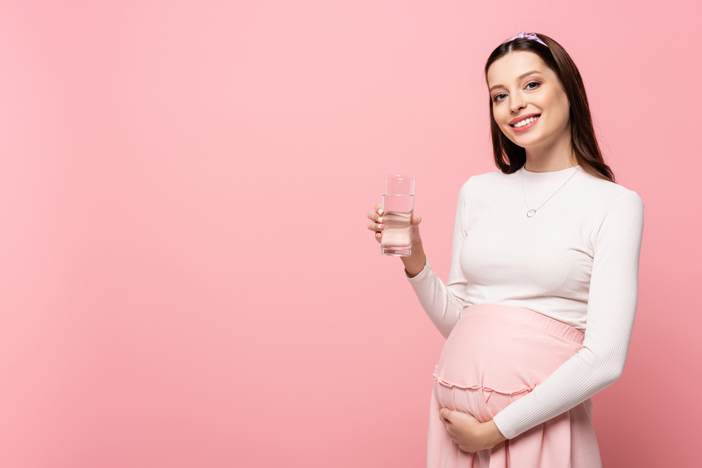 heureux jeune jolie femme enceinte avec verre d'eau isolé sur rose - Photo, image