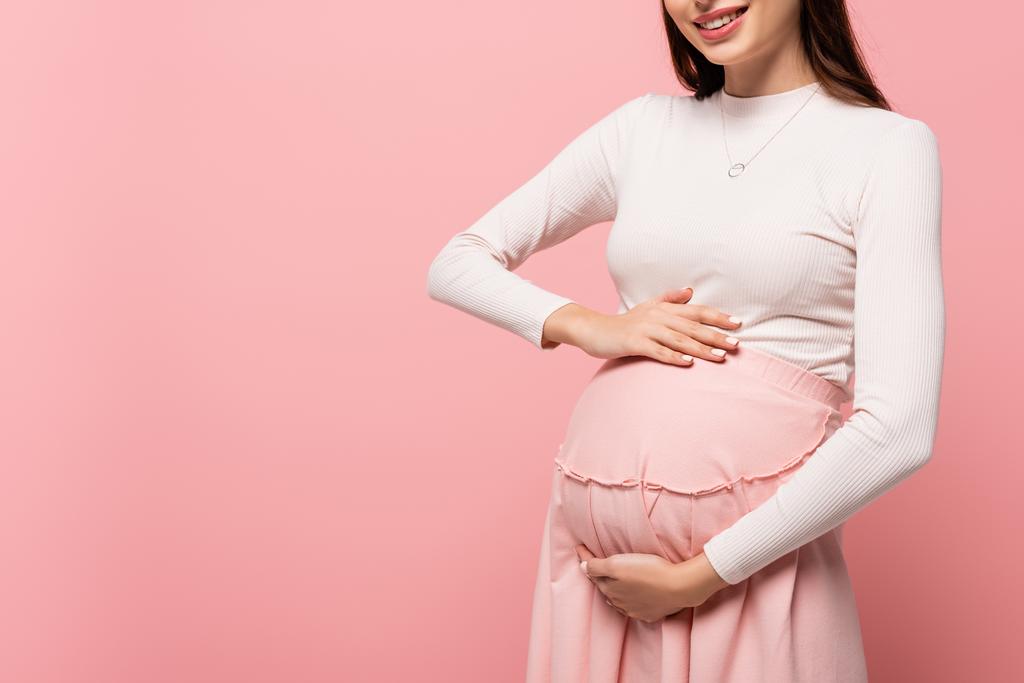 обрізаний вид щасливої молодої красивої вагітної жінки, яка торкається живота ізольовано на рожевому
 - Фото, зображення