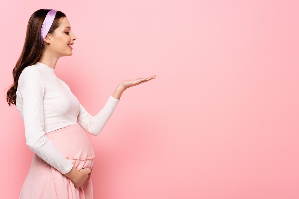 Ευτυχισμένη νεαρή όμορφη έγκυος γυναίκα που δείχνει με το χέρι απομονωμένο στο ροζ - Φωτογραφία, εικόνα