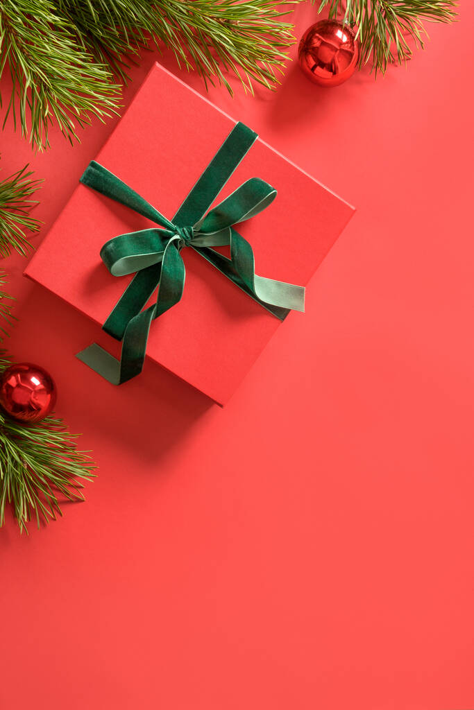 Різдвяний червоний подарунок з декоративною зеленою оксамитовою стрічкою, вічнозеленими гілками на червоній поверхні. Вид зверху. Вертикальна різдвяна листівка
. - Фото, зображення