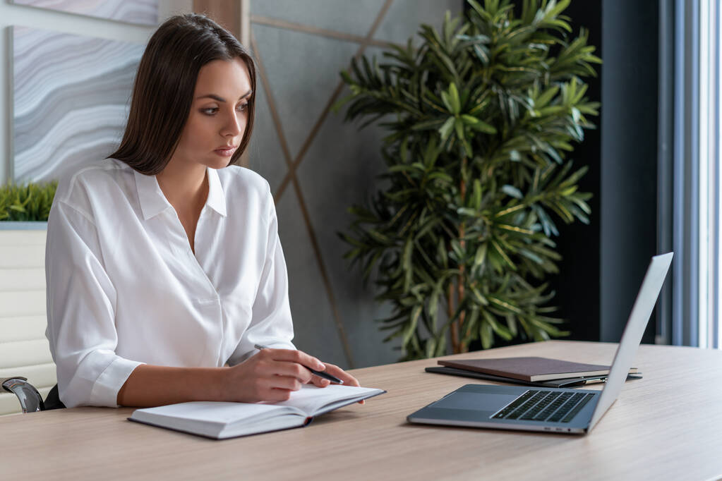 Kierownik biura kobieta w białej koszuli, siedząca przy stoliku z laptopem i notatnikiem, trzymająca długopis. Skoncentrowany pracownik biurowy patrzący na ekran w dużym, lekkim pokoju biurowym. Pojęcie pracy - Zdjęcie, obraz