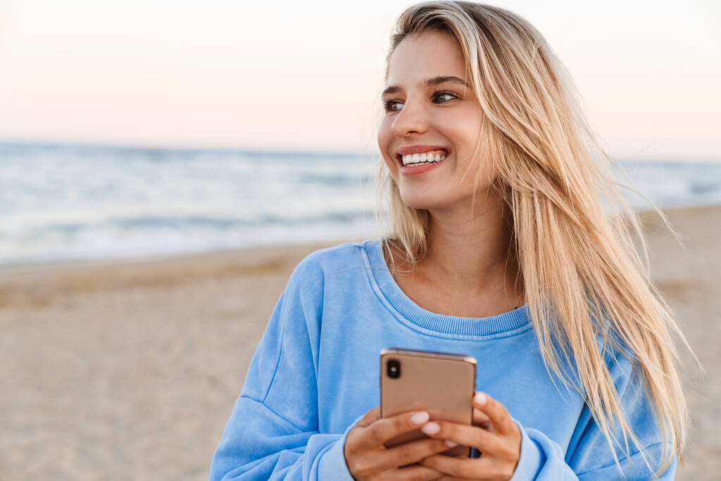 Nuori valkoihoinen blondi nainen hymyilee ja pitää matkapuhelinta kävellessään ulkona - Valokuva, kuva