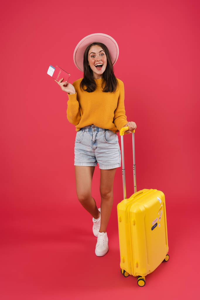 Volledige lengte van een vrolijke jonge vrouw die met de reistas loopt, met paspoort en tickets geïsoleerd over roze achtergrond - Foto, afbeelding