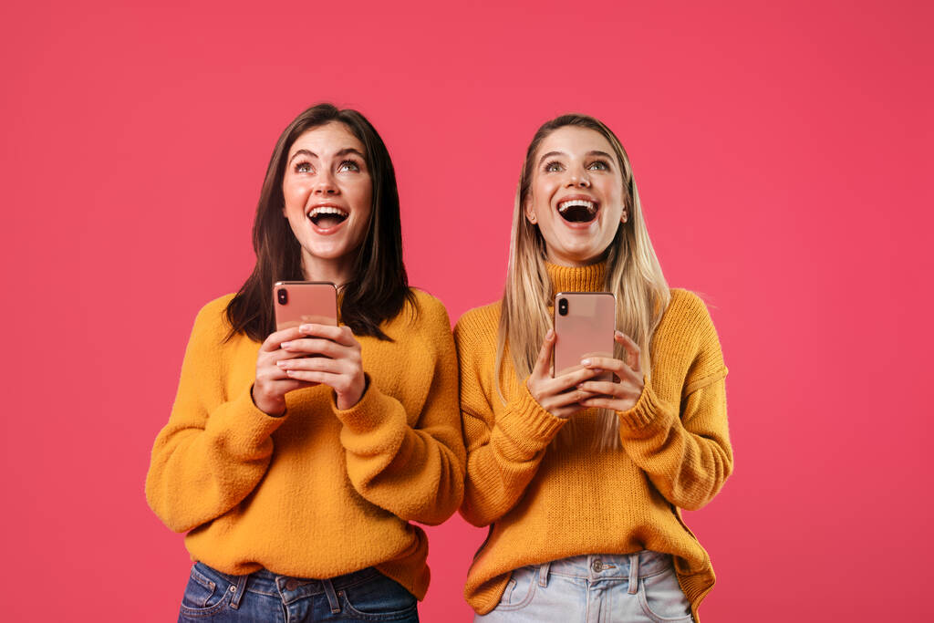 Изображение восхитительных милых женщин, улыбающихся при использовании смартфонов, изолированных на розовом фоне - Фото, изображение