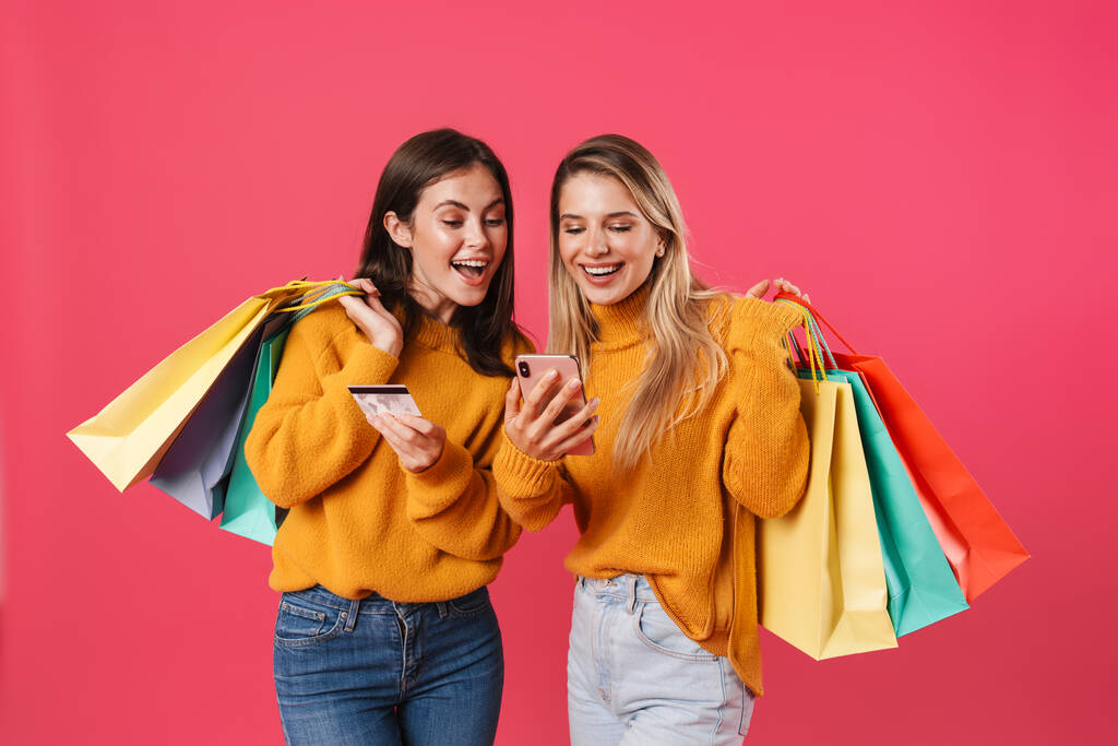Obrázek vzrušených žen držících kreditní kartu a mobil při pózování s nákupními taškami izolovanými přes růžovou stěnu - Fotografie, Obrázek