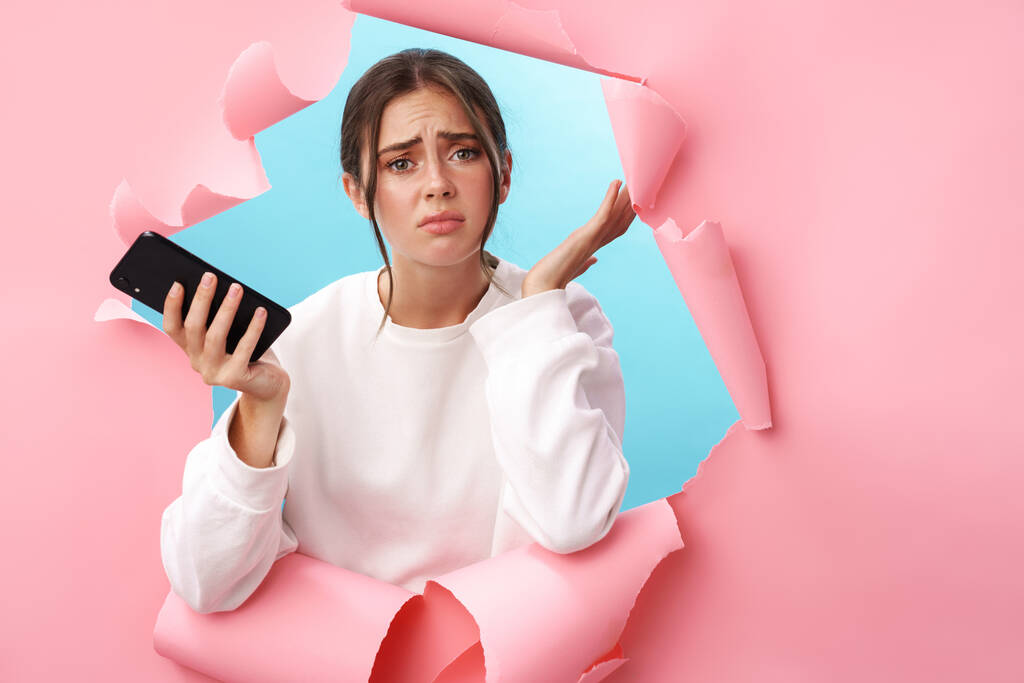 Caucasienne jeune femme exprimant la confusion tout en tenant le téléphone cellulaire isolé à travers fond rose - Photo, image