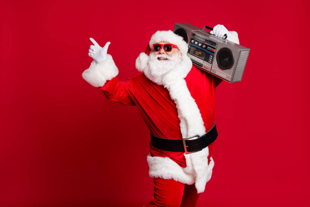 Hip-hop mucizesi. Emekli yaşlı bir adamın tam boy fotoğrafı gri sakallı gülüşü teyp kutusu boş parmaklı Noel Baba x-mas kostümlü güneş gözlüğü giysisi izole edilmiş kırmızı arka plan - Fotoğraf, Görsel