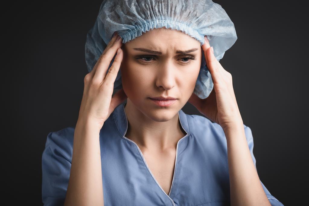 enfermera cansada en gorra médica que sufre de dolor de cabeza aislado en gris oscuro - Foto, imagen