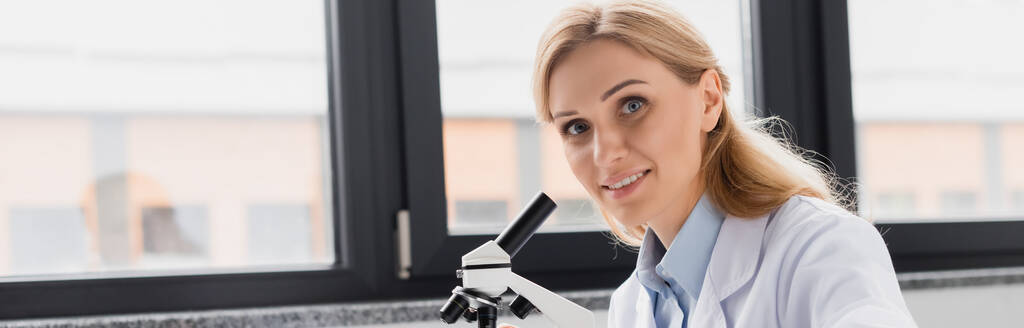 onnellinen tiedemies valkoisessa takissa katsomassa kameraa mikroskoopin lähellä laboratoriossa, banneri - Valokuva, kuva