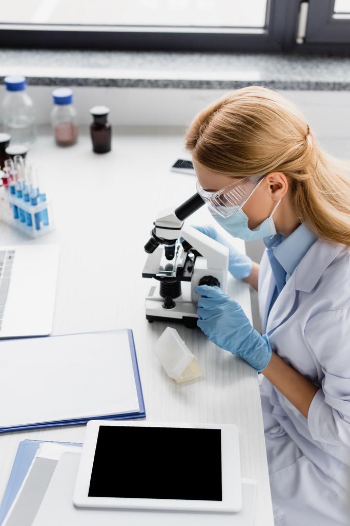 Wissenschaftler mit medizinischer Maske und Brille blickt durch das Mikroskop auf dem Schreibtisch - Foto, Bild