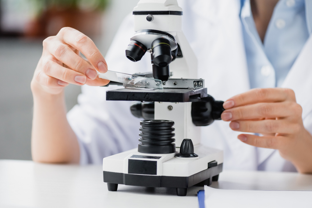 καλλιεργημένη άποψη της γυναίκας επιστήμονα που κρατά γυάλινη πλάκα δοκιμής κοντά στο μικροσκόπιο στο εργαστήριο - Φωτογραφία, εικόνα