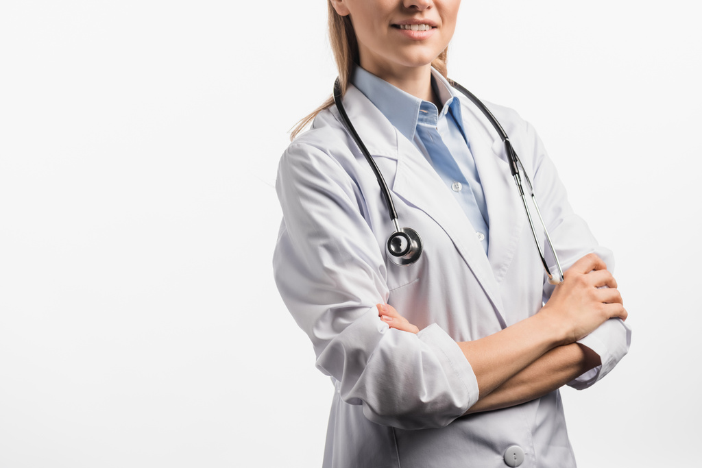 visão recortada de enfermeira alegre em casaco branco de pé com braços cruzados isolados em branco - Foto, Imagem